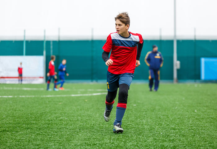 A young boy wearing a red and blue soccer kit is sprinting on a green artificial turf field during practice.