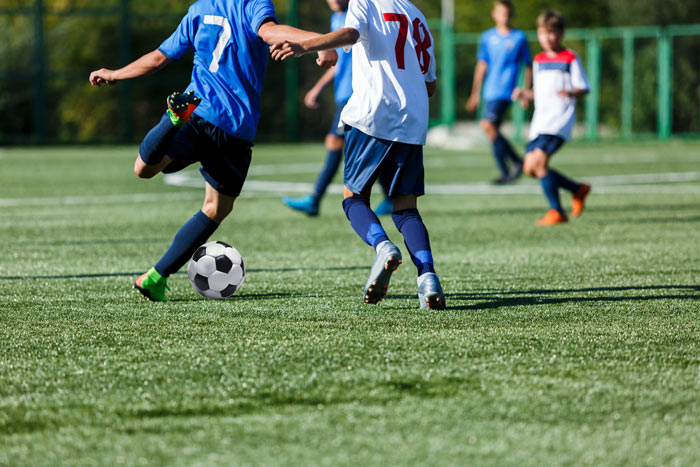 Two young soccer players on a grassy field, one in a blue jersey kicking a black and white soccer ball, while another player in a white jersey follows closely. A soccer match in progress.