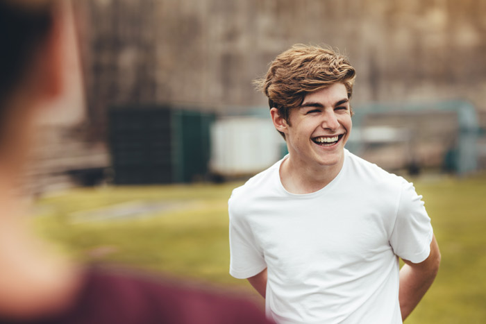 A young man with a joyful smile, wearing a white t-shirt, laughing in an outdoor setting.