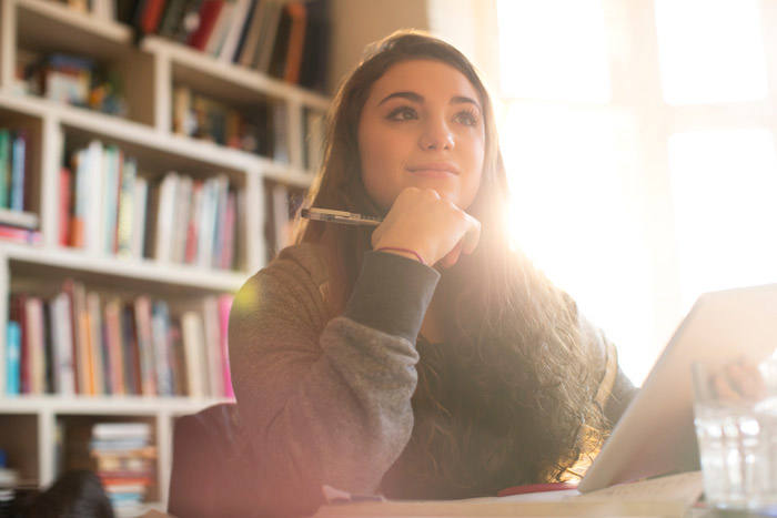 A young woman sitting at a desk, thoughtfully resting her chin on her hand while holding a pen, with a stack of books and sunlight streaming through a window in the background. Ideal for capturing a moment of study or reflection.