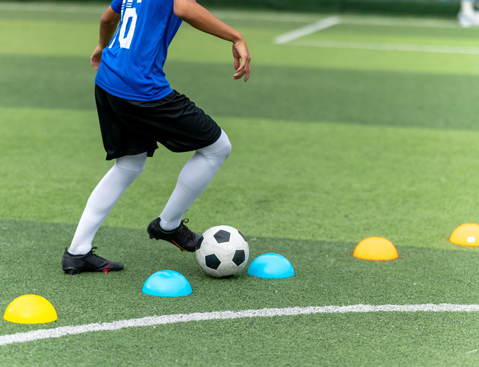 A young soccer player dribbles a ball past colorful training cones on a green field, wearing a blue jersey and black shorts.